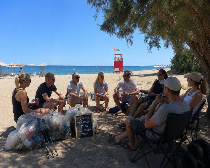 A group of people sitting in folding chairs on a sandy beach under a tree, gathered around trash bags and a small sign. The background shows the blue ocean, beach umbrellas, and a red lifeguard tower.